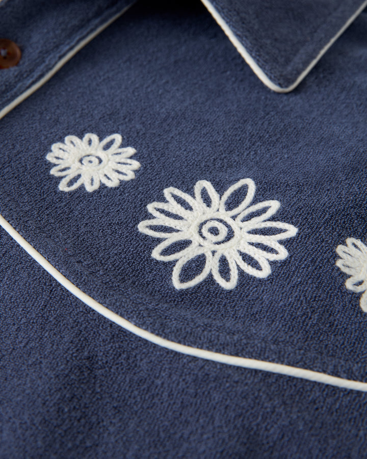 A close-up of The Gaucho Shirt - Moontide by Dandy Del Mar shows blue fabric with white floral embroidery and piping, echoing the classic Gaucho Shirt style.