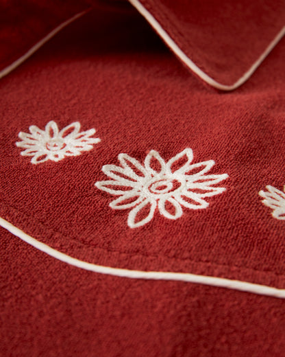 A close-up of The Gaucho Shirt - Rouge by Dandy Del Mar showcases red fabric with white embroidered floral patterns and white piping.