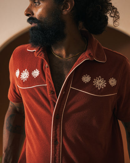 A person with a beard wears Dandy Del Mar's rust-colored Gaucho Short - Rouge featuring floral embroidery and an elastic waistband while looking to the side against a softly lit background.