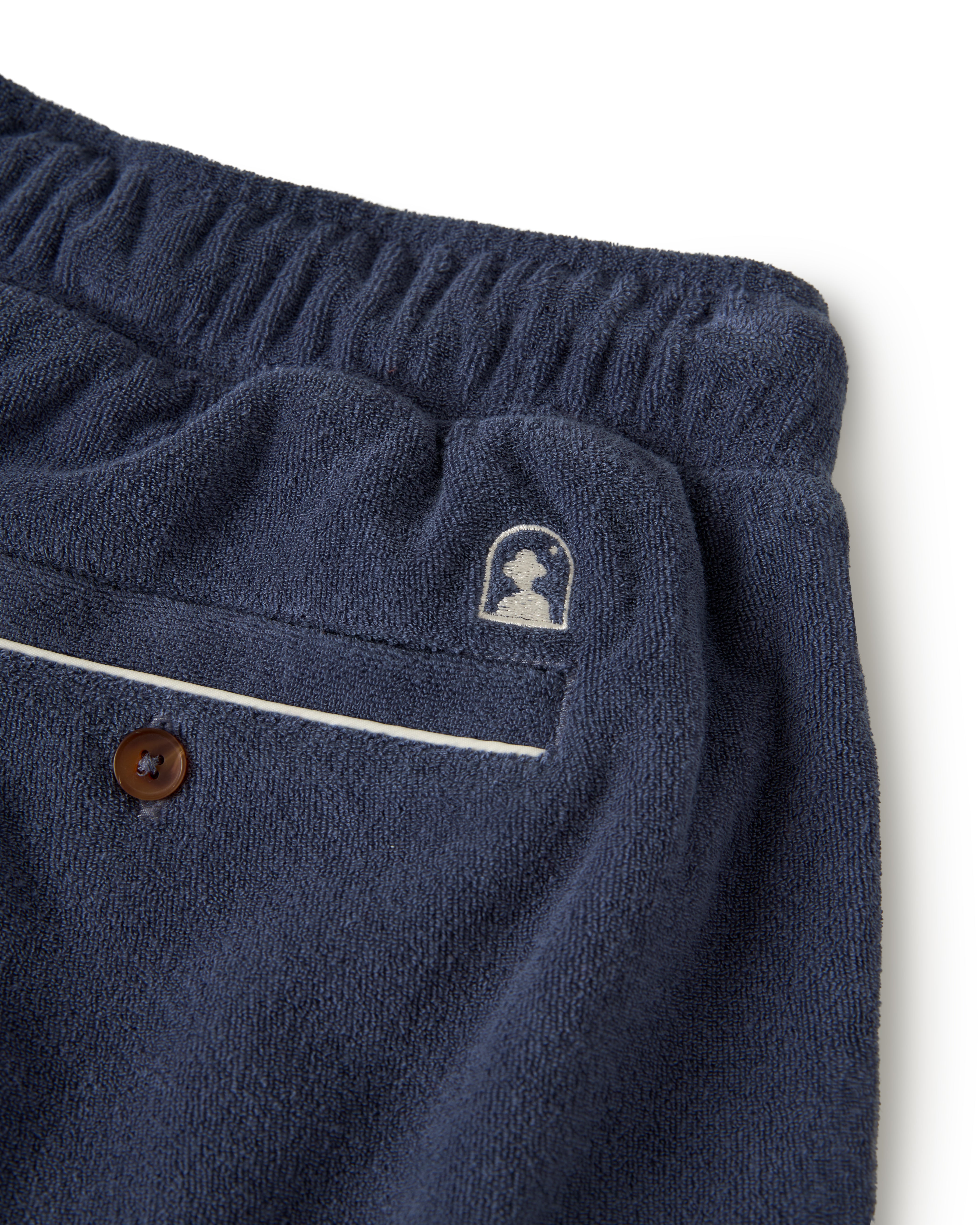 Close-up of The Gaucho Short - Moontide by Dandy Del Mar: navy blue pants with an elastic waistband, a back pocket with embroidered logo, and a brown button.