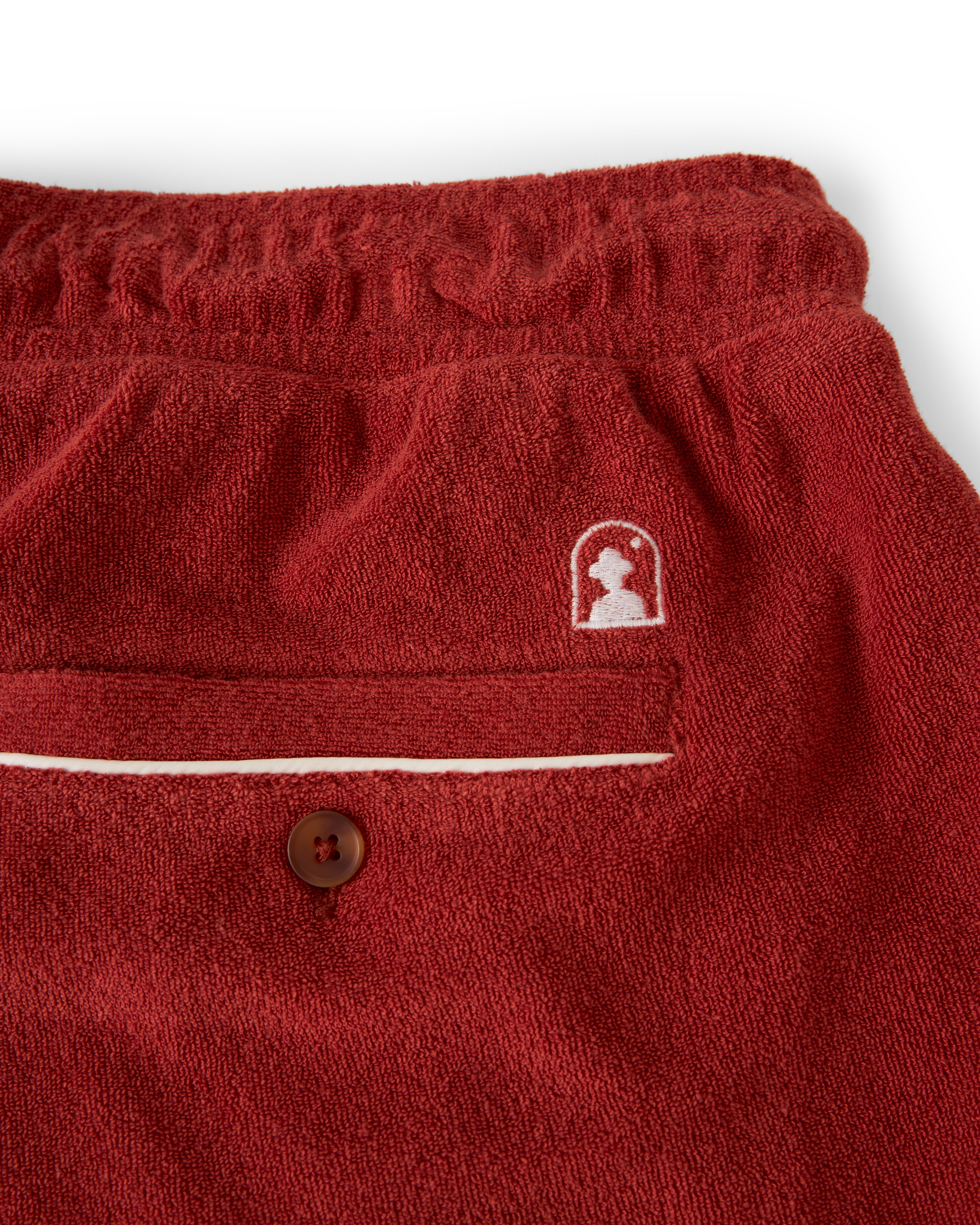 Close-up of Dandy Del Mar's The Gaucho Short - Rouge in terry cloth with an embroidered logo above a buttoned pocket and an elastic waistband.