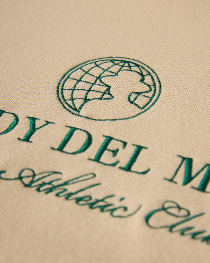 Close-up of The Marseille French Terry Pullover - Sabbia by Dandy Del Mar, showcasing a green logo with a globe and lion silhouette, alongside partial text "DY DEL M... Athletic Club" in stylized font, reminiscent of Raglan Pullover design with Dandy Del Mar chest embroidery.