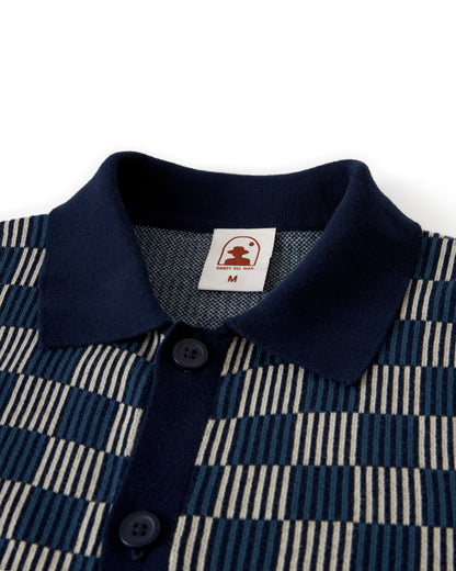 Close-up of The Martinique Sweater-Knit Shirt - Anchor by Dandy Del Mar featuring a checkered blue and beige pattern, collared design with buttons, and a visible tag displaying a small logo and size information, showcasing its iconic check-knit motif.