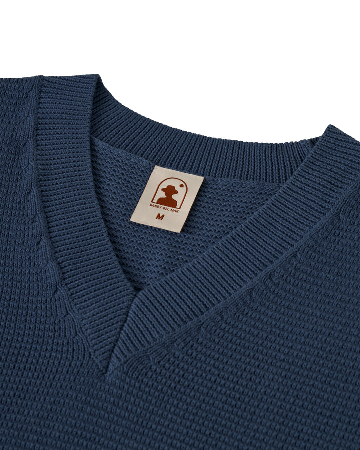 Close-up of The Montego Knit Vest in Moontide by Dandy Del Mar, featuring a visible label with a chess piece logo and size 'M.'.