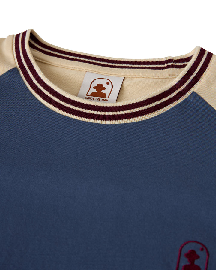 The Nassau Tee by Dandy Del Mar, in blue and cream cotton jersey, sports a crew neck with maroon trim and an embroidered logo on the front and inside tag.