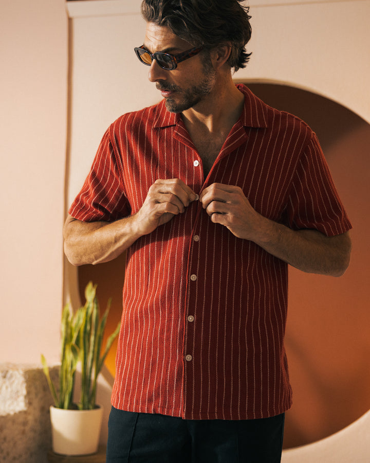 A man with sunglasses is buttoning a red, short-sleeve, striped Palma Shirt - Garam from Dandy Del Mar with a camp collar. He is posed in front of a wall with a circular cutout and a plant to his side.