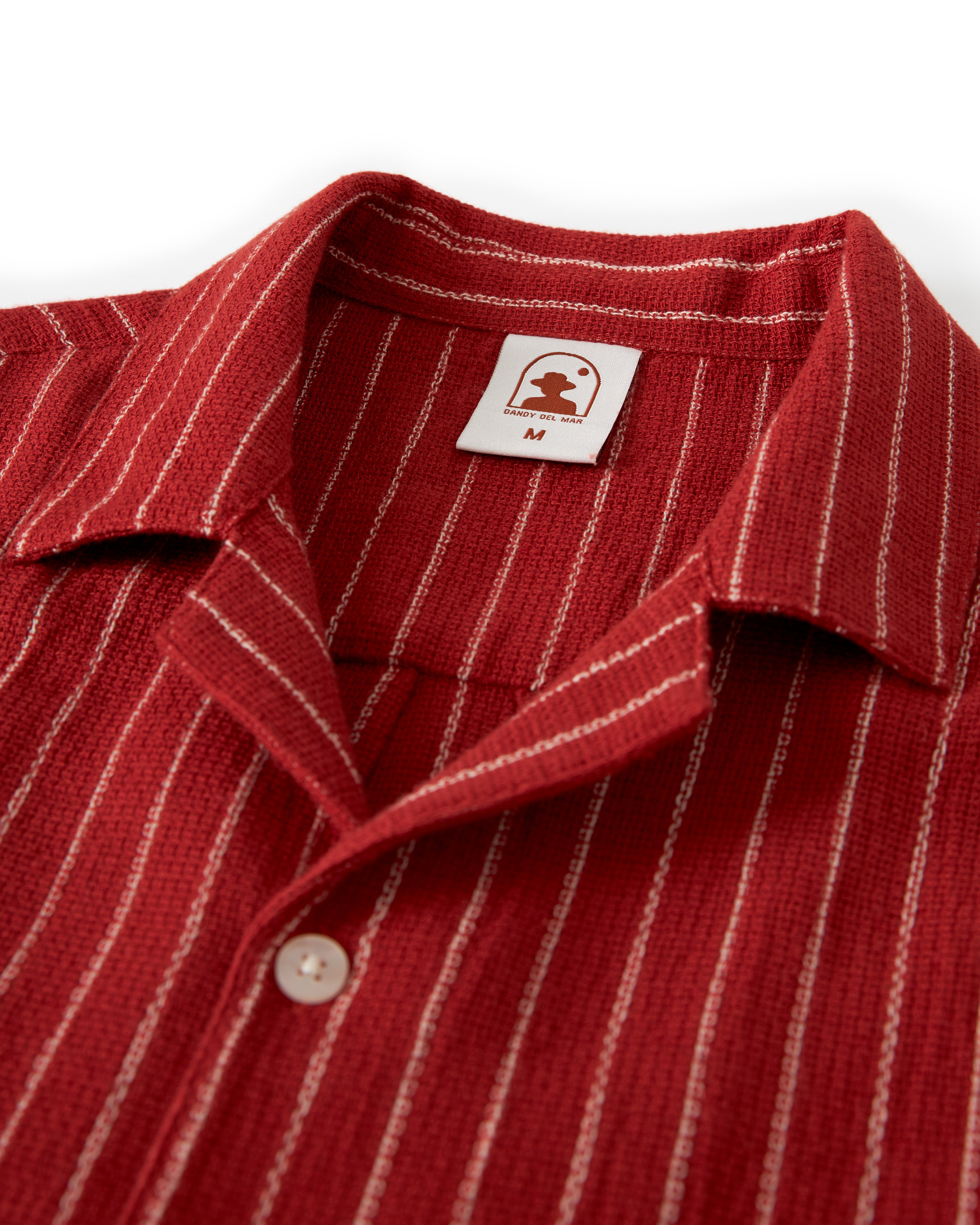 Close-up of The Palma Shirt - Garam by Dandy Del Mar, showcasing its red fabric with white vertical stripes, including a sewn-on label at the back of the camp collar featuring a small logo and the letter 'M'.