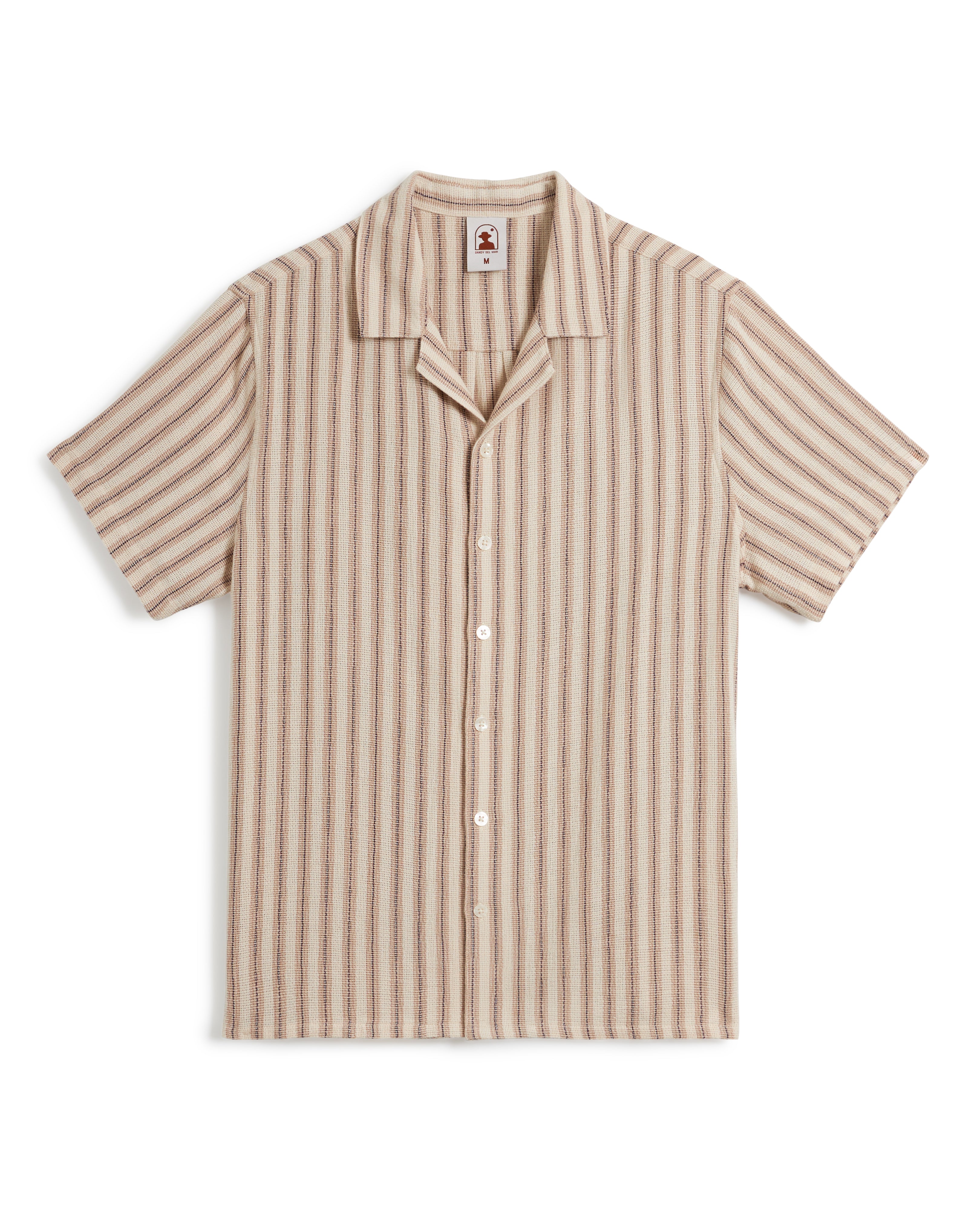 The Palma Shirt - Scallop by Dandy Del Mar is a short-sleeve shirt with a camp collar, showcasing a beige base and yarn-dyed stripe canvas pattern in various shades of brown. It features five white buttons down the front.