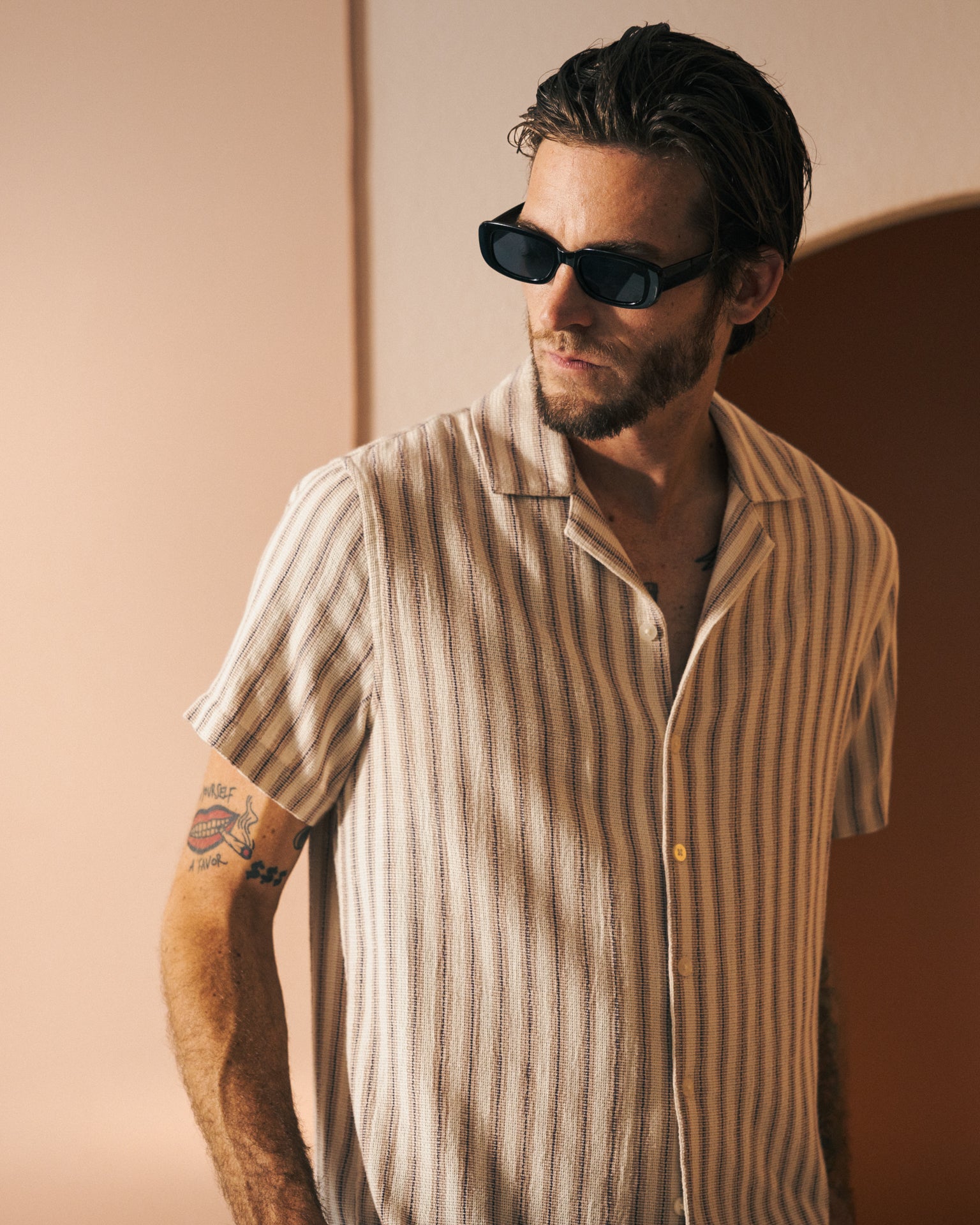 A man with sunglasses and a beard stands in a sunlit room, wearing The Palma Shirt - Scallop from Dandy Del Mar. Tattoos are visible on his left arm, adding to his distinctive style that evokes the feel of exotic spices.