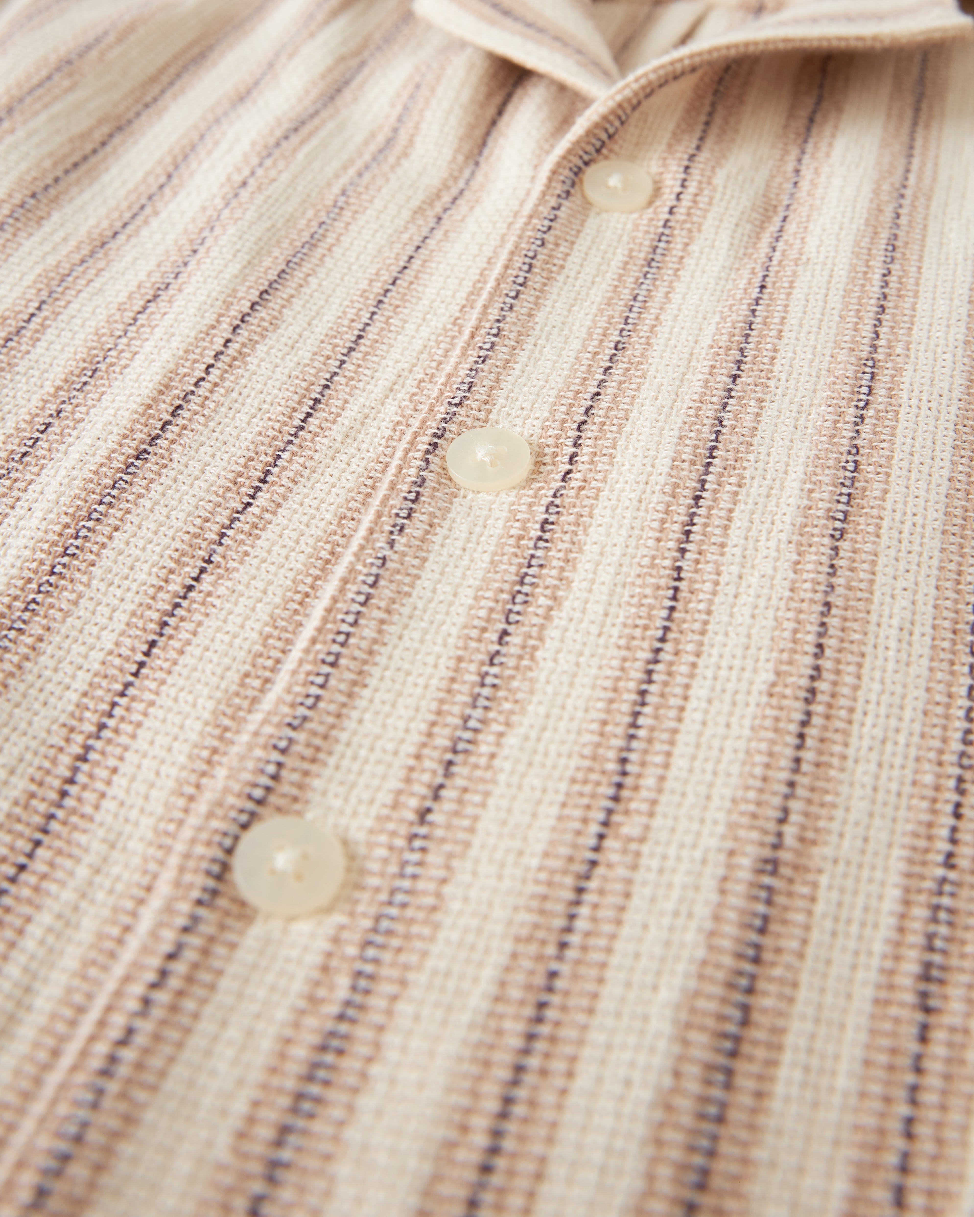 A close-up image of The Palma Shirt - Scallop by Dandy Del Mar, showcasing its beige and brown striped pattern with three visible white buttons, crafted from soft burlap-textured fabric.