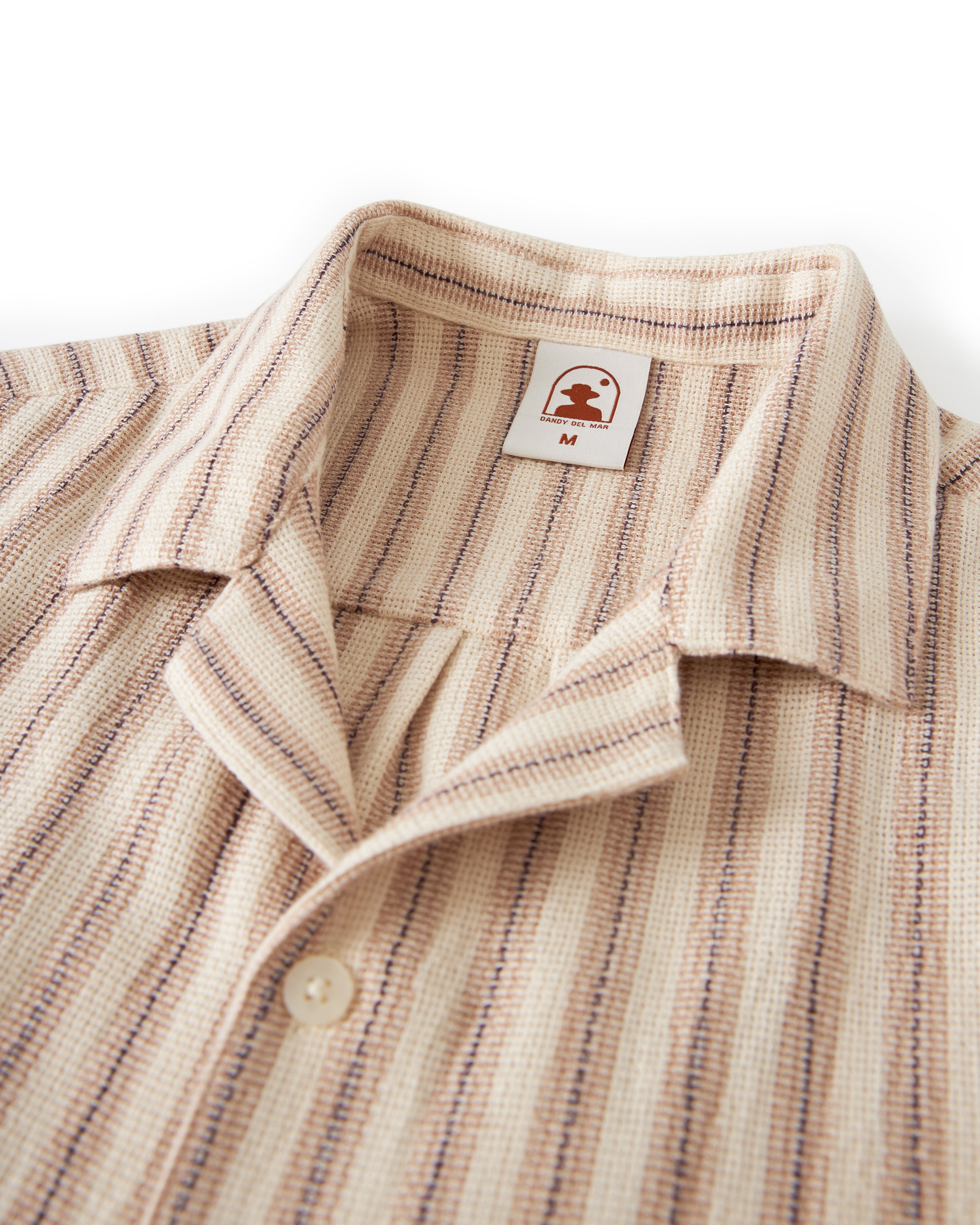 Close-up of The Palma Shirt - Scallop by Dandy Del Mar, a beige shirt featuring vertical brown and blue stripes, a camp collar, and two buttons. Made from soft burlap-textured fabric, it displays a label with a small logo and the size "M.