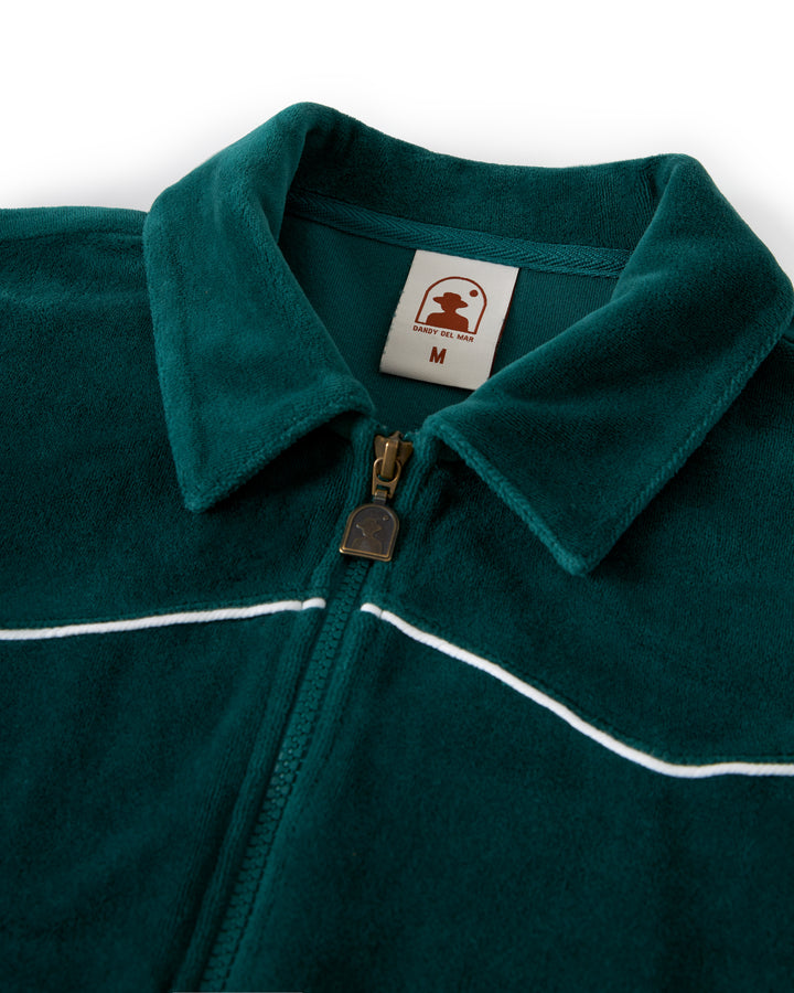 Close-up of The Rio Velour Jacket - Albero by Dandy Del Mar, a dark green zip-up fleece jacket with a white-accented collar and the label "M" for medium. The label features a red logo of a person wearing a hat. This design exudes an athleticism that sets it apart, reminiscent of classic velour track jackets.