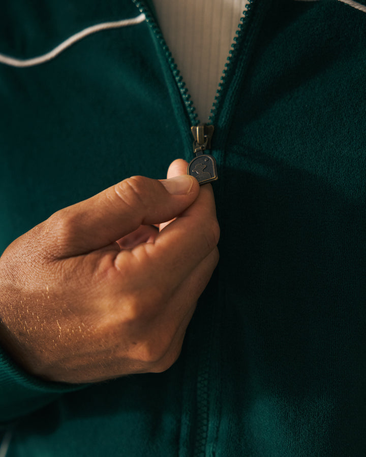 A person is shown zipping up The Rio Velour Jacket - Albero by Dandy Del Mar, focusing on their hand gripping the zipper pull.