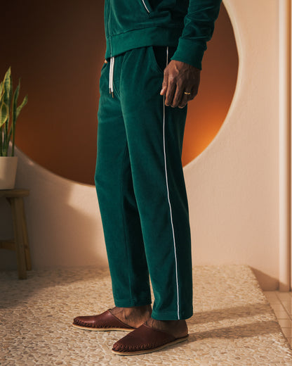 A person wearing The Rio Velour Pant - Albero by Dandy Del Mar, paired with brown slippers, stands beside a wooden stool holding a potted plant in front of a round, backlit wall, effortlessly blending leisure and athletics.