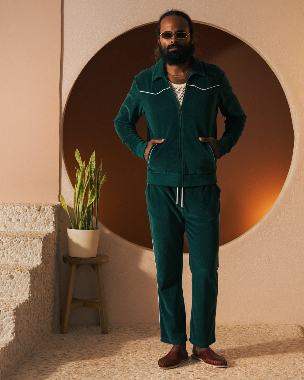 A person with a beard and sunglasses stands in front of a circular backdrop, wearing The Rio Velour Pant - Albero from Dandy Del Mar paired with brown shoes. A potted plant is placed on a stool to the left.