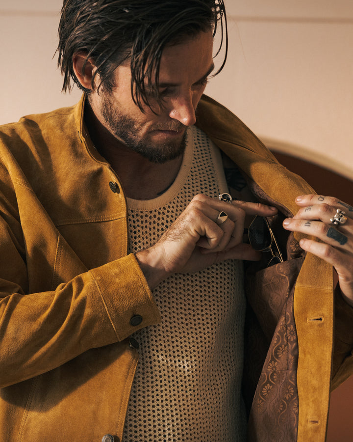 A man in "The San Juan Suede Leather Jacket - Sable" by Dandy Del Mar, featuring brass buttons, inspects the inside pocket while holding a pair of sunglasses. He is wearing a white mesh top and silver rings.