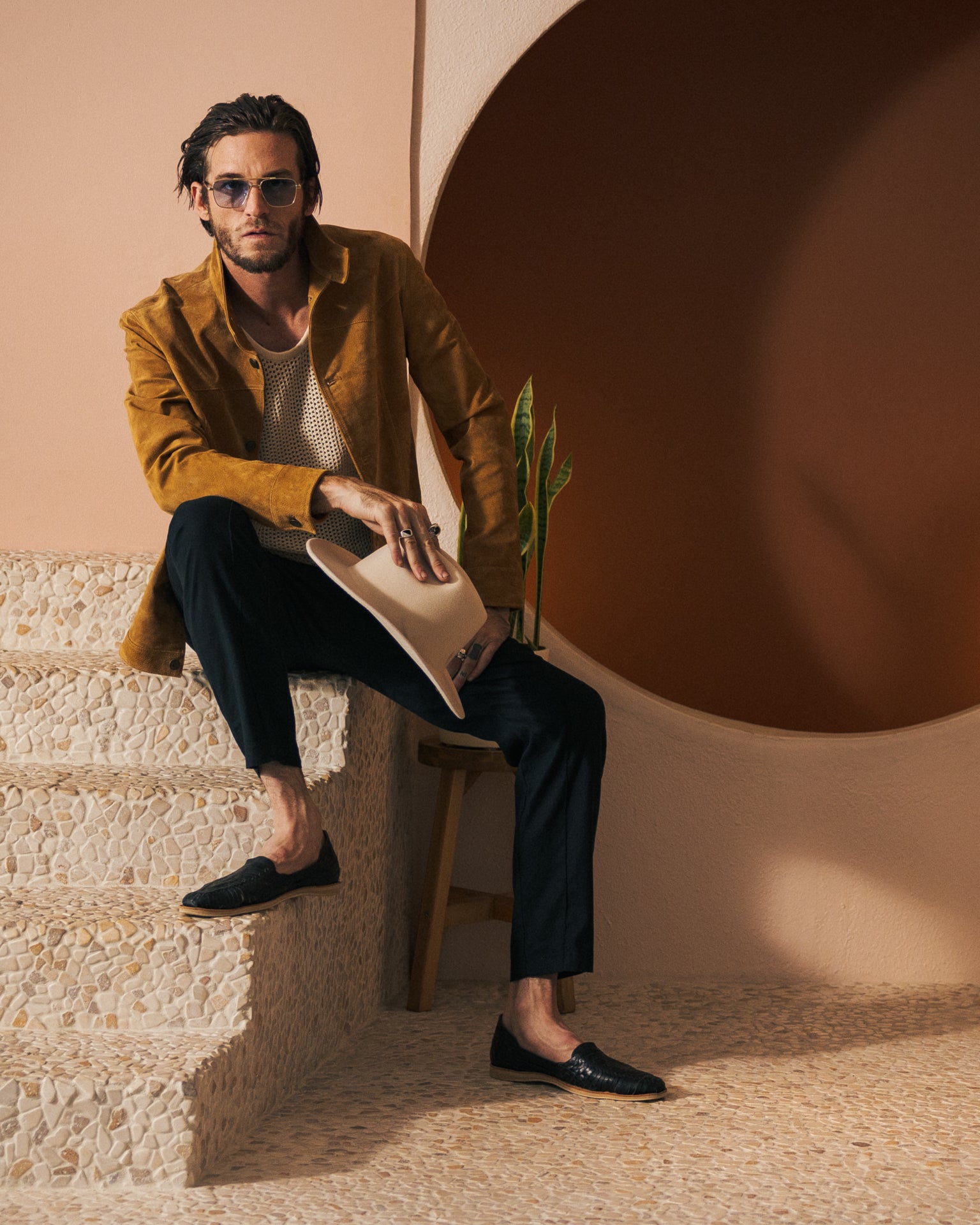 A man in The San Juan Suede Leather Jacket - Sable by Dandy Del Mar, paired with black pants, sits on stairs holding a hat. In the background, there is a plant and a brown circular wall feature.