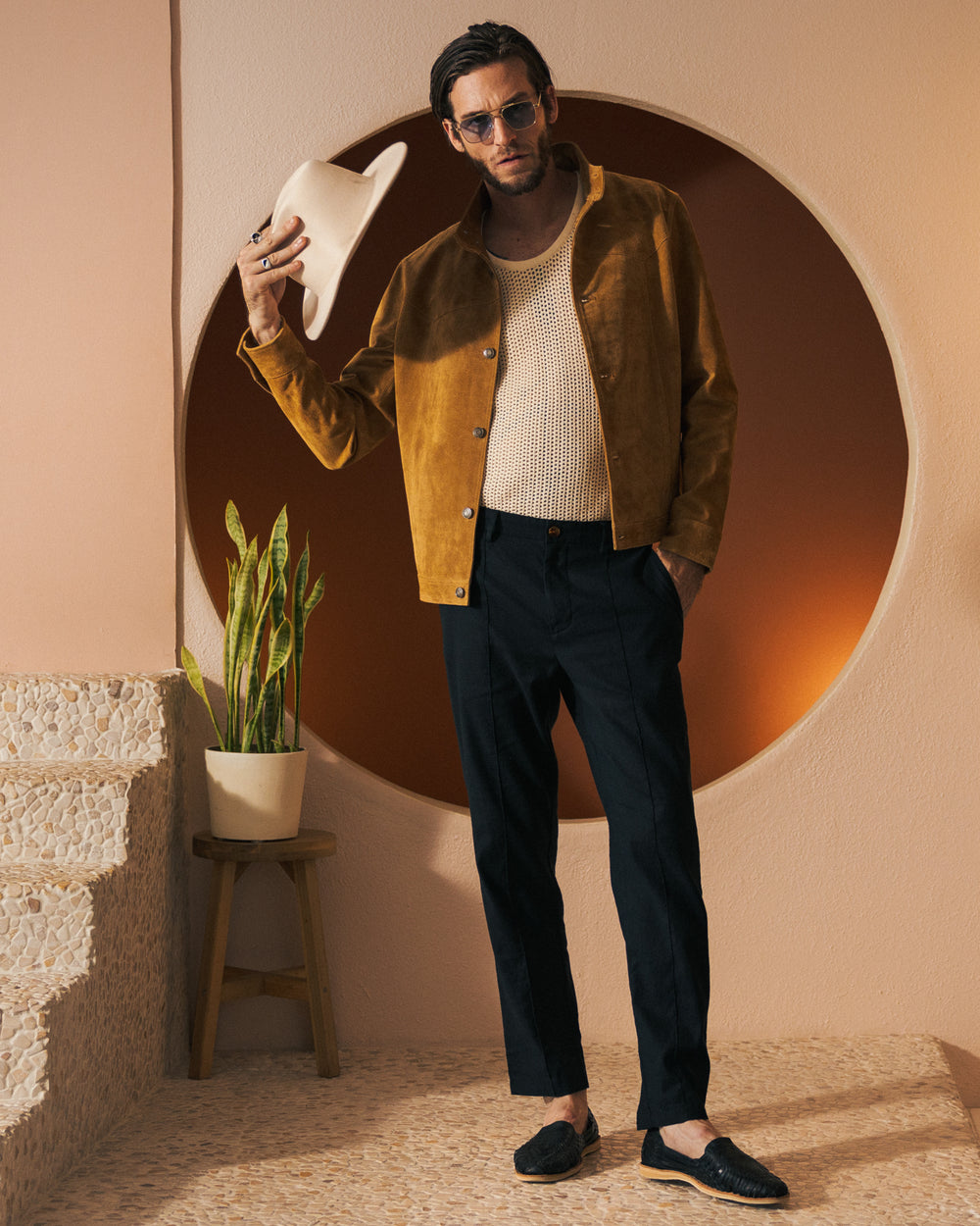 A person wearing sunglasses, a Dandy Del Mar "The San Juan Suede Leather Jacket - Sable," a white shirt, and black pants holds a white hat in their right hand while standing next to a plant on a stool in a stylish indoor setting.