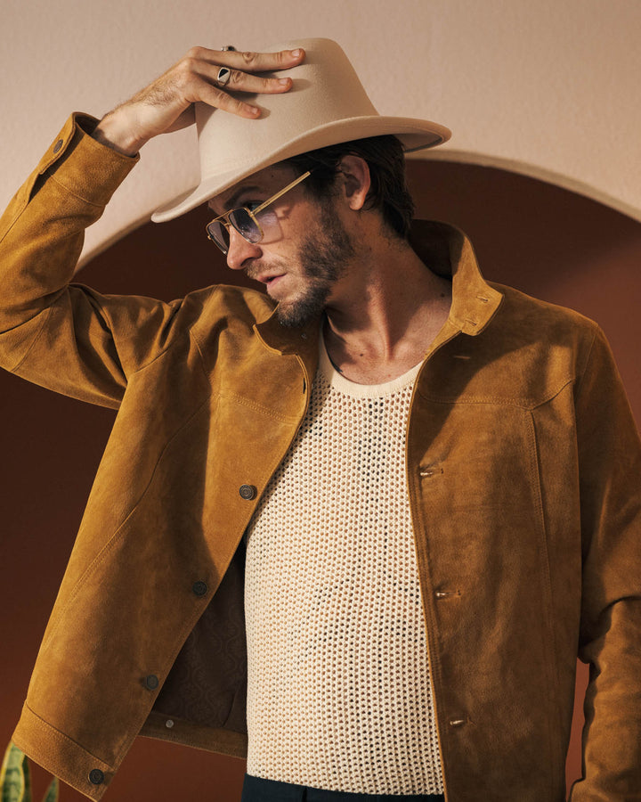 A man wearing The San Juan Suede Leather Jacket - Sable by Dandy Del Mar, a white mesh tank top, and a beige hat touches the brim of his hat while standing in front of an archway.