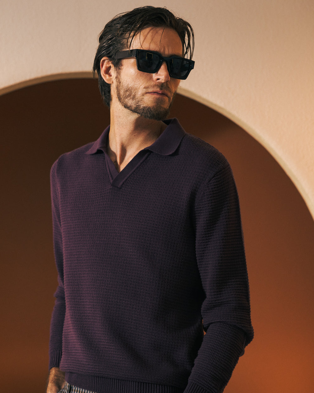 A man with dark hair and a beard wears black sunglasses and Dandy Del Mar's "The Sebastian Long Sleeve" in Amethyst, standing in front of a light brown background with a textured finish.