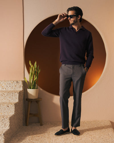 A man in sunglasses wears the Dandy Del Mar Sebastian LS Polo Shirt in Amethyst and patterned pants, standing by a potted plant and stairs, with a circular wall cutout behind him.