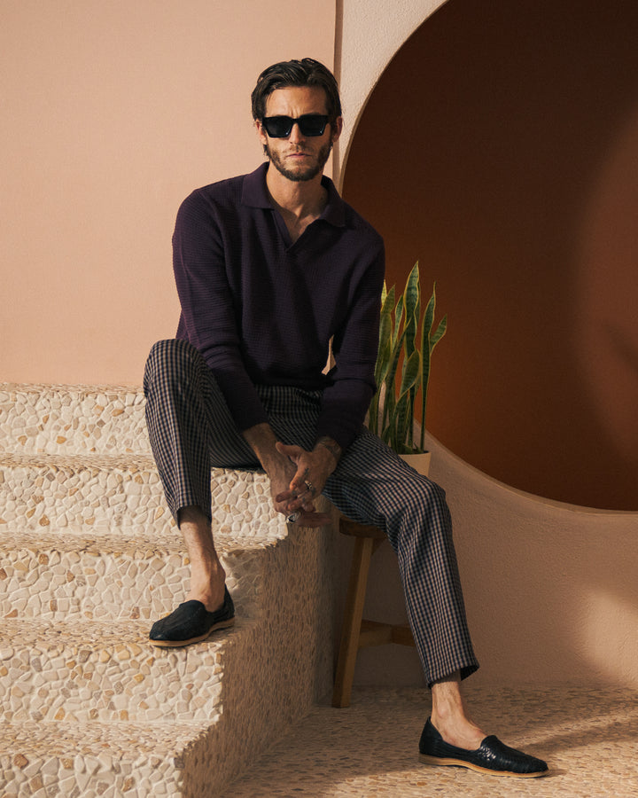 A man wearing dark sunglasses, an Amethyst-colored The Sebastian Long Sleeve by Dandy Del Mar, and checkered pants is sitting on a pebble-textured staircase next to a small potted plant.