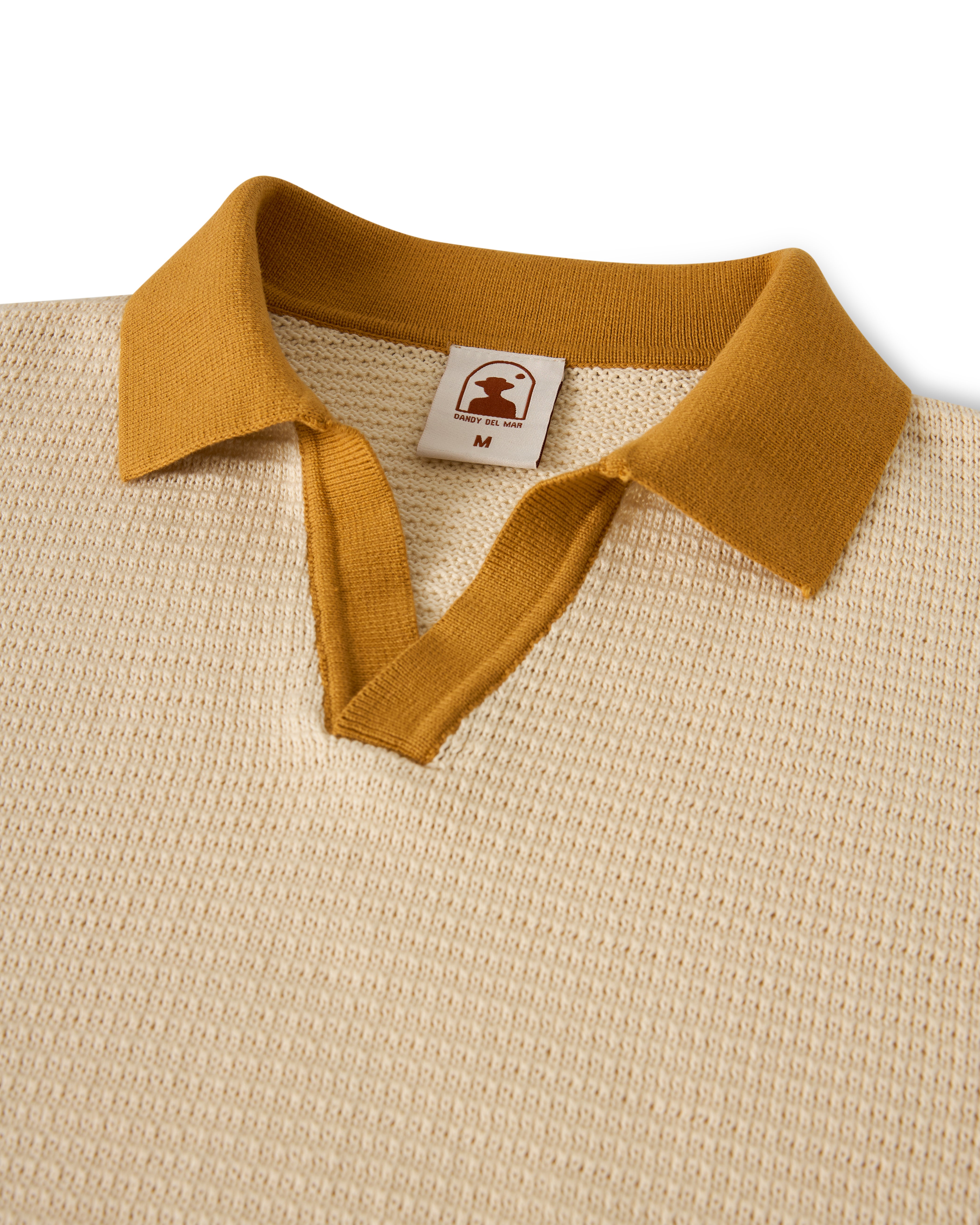 The Sebastian Long Sleeve Polo by Dandy Del Mar in Vintage Ivory is a beige sweater-knit cotton polo shirt featuring a mustard collar and placket, textured finish, and a brown logo tag with a bell. Size "M" is marked inside the collar.