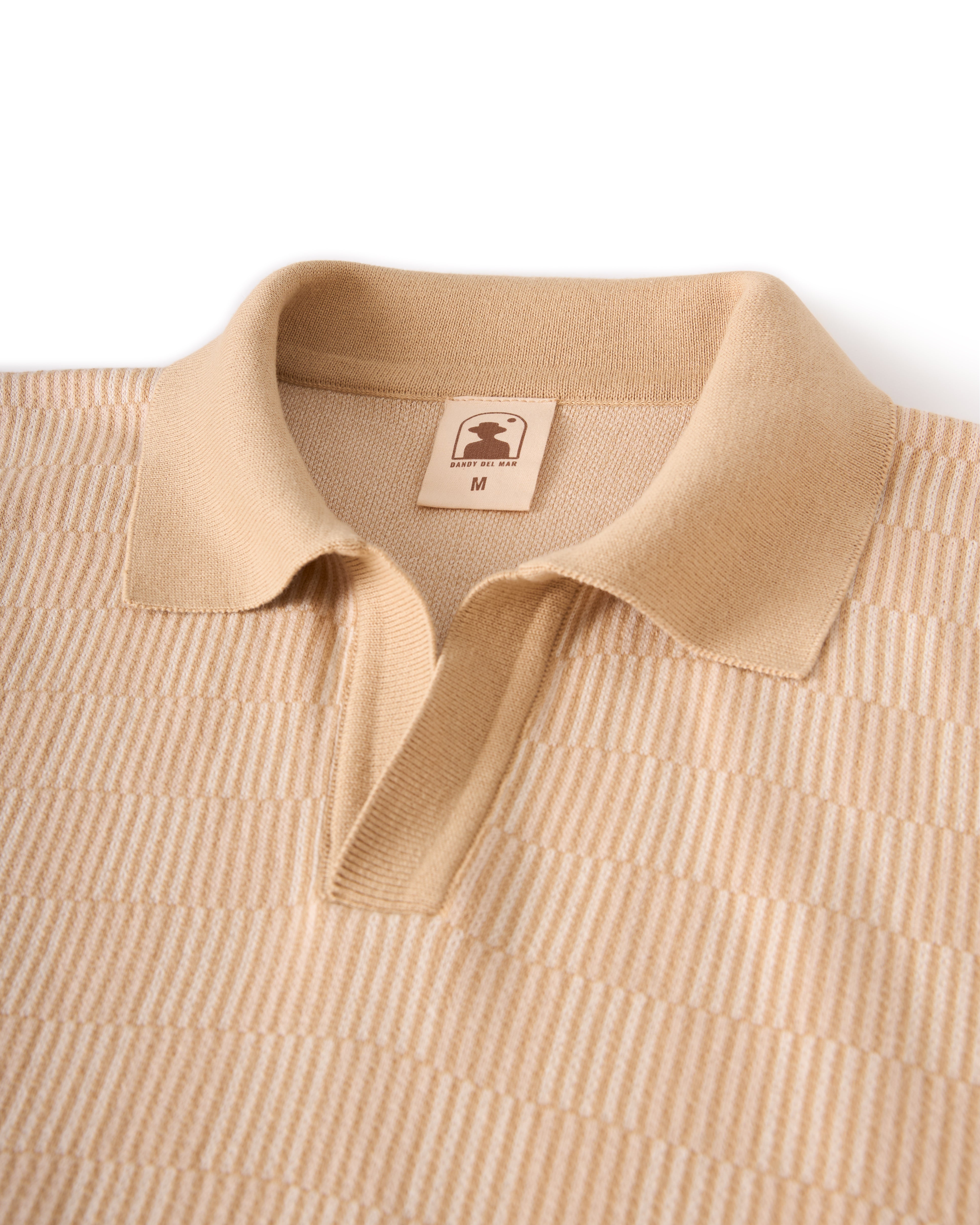 The Sebastian Knit Polo - Shell by Dandy Del Mar is a beige knit collared shirt with a medium size tag, featuring a textured stripe pattern reminiscent of classic Italian knitwear.