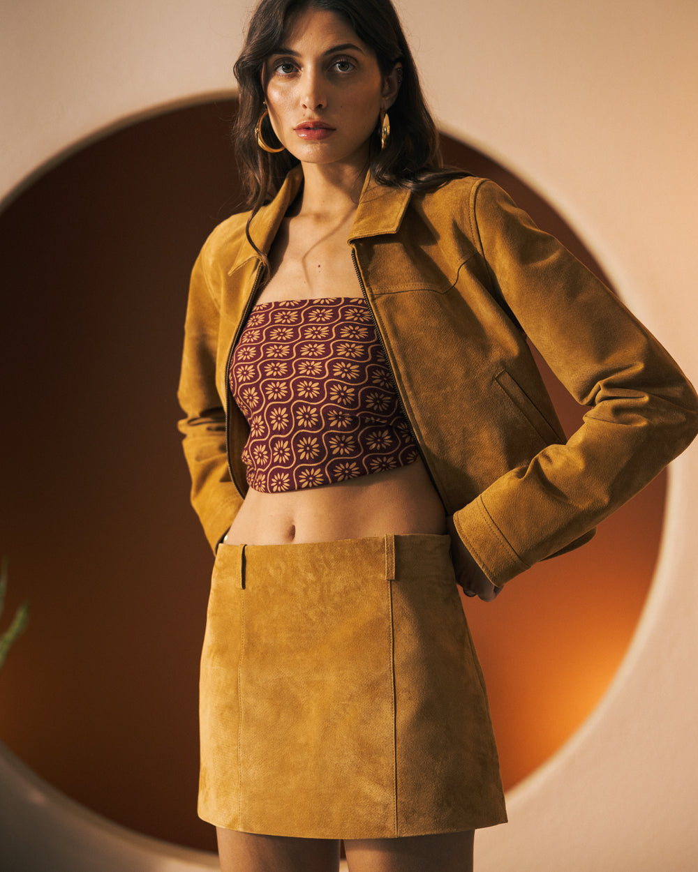 A woman stands in front of a circular backdrop, wearing a mustard-colored jacket paired with a patterned strapless top and The Sicily Leather Skirt - Sable by Dandy Del Mar.