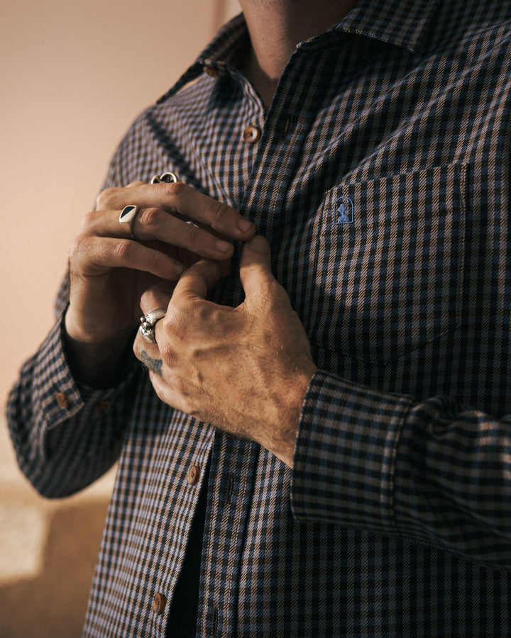 A person wearing a long-sleeve, houndstooth Tresco Shirt from Dandy Del Mar is buttoning it up with casual elegance. The tailored fit highlights their style, and several rings adorn their fingers.