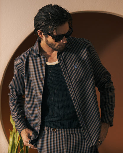 A man with slicked-back hair exudes casual elegance, wearing sunglasses and a finely tailored outfit—a Tierra Houndstooth Tresco Shirt from Dandy Del Mar paired with a black knit top and plaid pants—while standing in front of an archway.