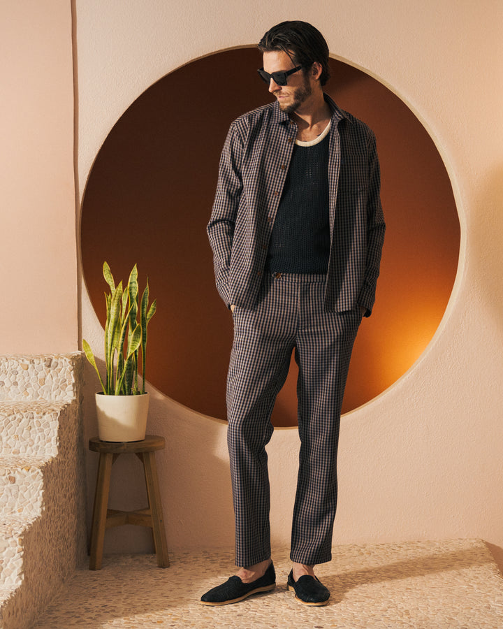 A man dressed in a checkered suit and the black Tierra Houndstooth Tresco Shirt from Dandy Del Mar, exuding casual elegance, stands in front of a circular orange backdrop. A potted plant is placed beside him on steps.