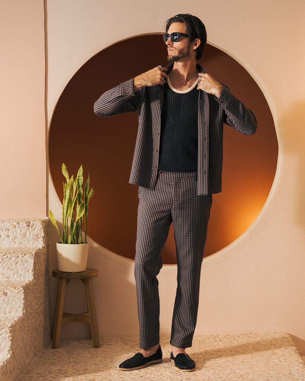 A man in a checked suit jacket and tailored fit trousers stands on a textured staircase near a potted plant, adjusting his jacket. With an air of casual elegance, he sports sunglasses and the The Tresco Shirt - Tierra Houndstooth by Dandy Del Mar.