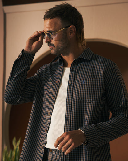 A man in a checkered shirt and white undershirt, reminiscent of The Tresco Trouser - Tierra Houndstooth pattern by Dandy Del Mar, adjusts his sunglasses while standing indoors.