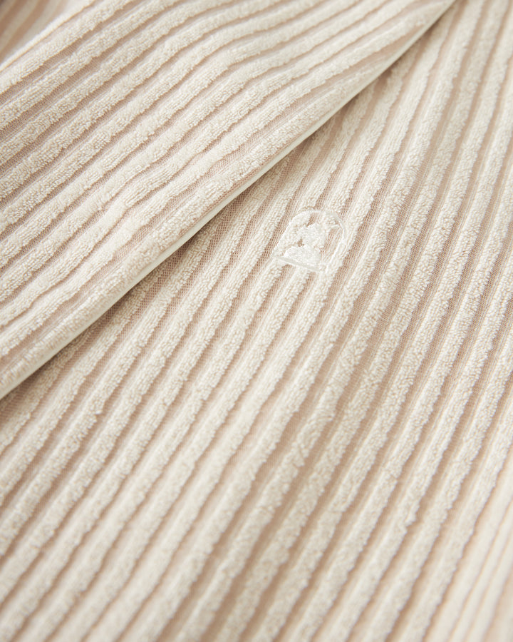 Close-up of beige, ribbed fabric with an embossed Dandy Del Mar logo measuring approximately 1-inch square detail near the center, showcasing the sophisticated jacquard scallop stripe texture of The Tropez Jacquard Robe.