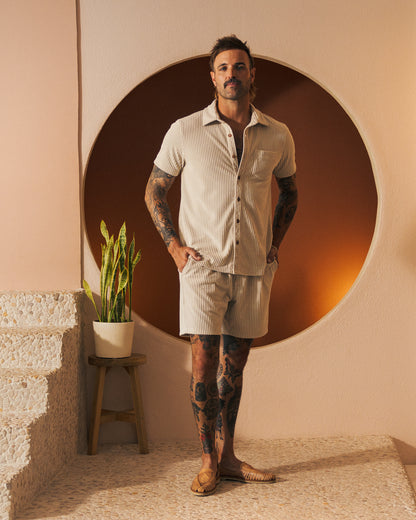 A man with tattoos stands near a potted plant on a stool, wearing Dandy Del Mar's The Tropez Jacquard Short - Scallop Stripe with an elastic waistband and a matching short-sleeved shirt. He is positioned in front of a circular wall feature.