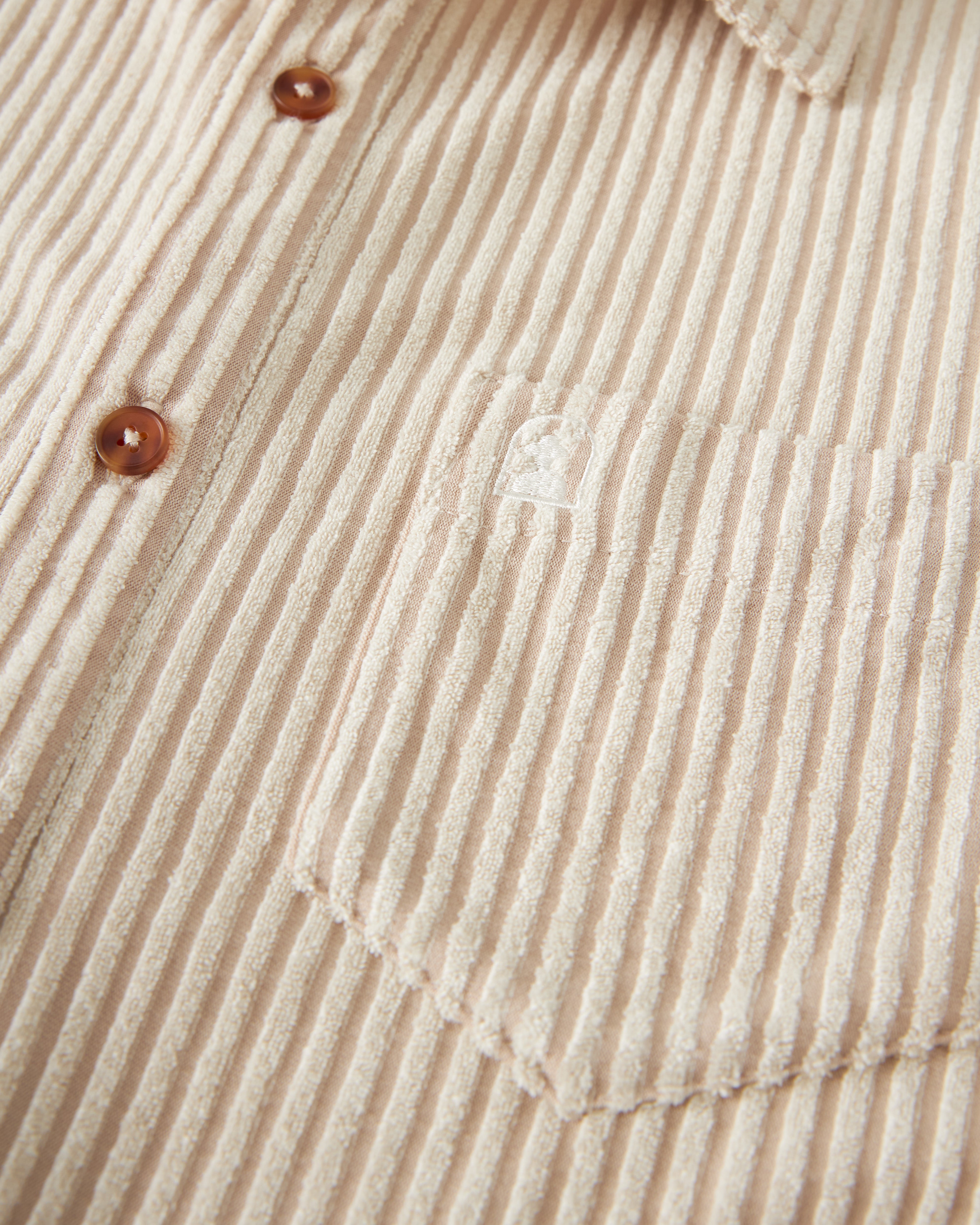 Close-up of a beige corduroy Tropez Jacquard Shirt - Scallop Stripe by Dandy Del Mar, featuring brown buttons and an embroidered logo on the chest pocket, reminiscent of the Côte d'Azur's effortless style.