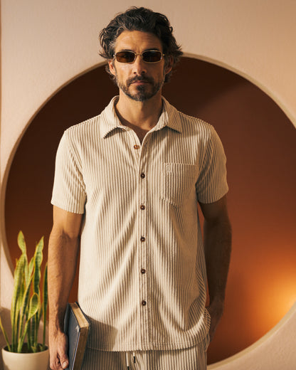 A man with short wavy hair and a beard, wearing sunglasses and a beige Tropez Jacquard Shirt in Scallop Stripe from Dandy Del Mar, holds a book. A plant and a round wall feature are in the background, evoking the effortless charm of Côte d'Azur.