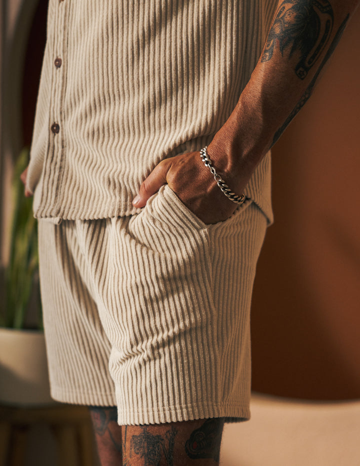 Close-up of a person wearing The Tropez Jacquard Short - Scallop Stripe by Dandy Del Mar, paired with a matching beige jacquard striped terry button-up shirt. One hand is tucked into the elastic waistband pocket, revealing their tattoos and a silver bracelet on their wrist.