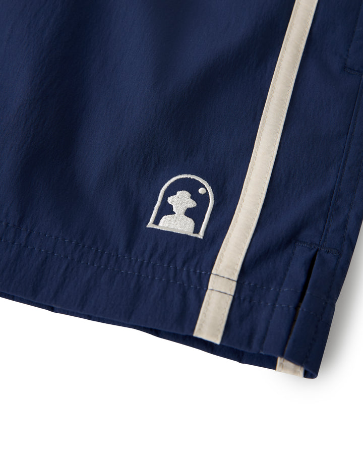 Close-up of Dandy Del Mar's The Ventura Volley Short - Anchor featuring navy blue swim shorts with a white embroidered figure-under-arch logo, beige side stripes, an elastic waistband, and a micro mesh liner for comfort.