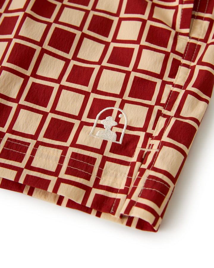 Close-up of fabric from The Ventura Volley Short - Rouge by Dandy Del Mar, showcasing red and tan geometric squares with a white embroidered emblem and a small figure under an arch.