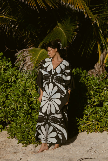A woman in The Tropea Kaftan - Albatross by Dandy Del Mar stands barefoot on sand near green bushes and palm trees, looking to the side.