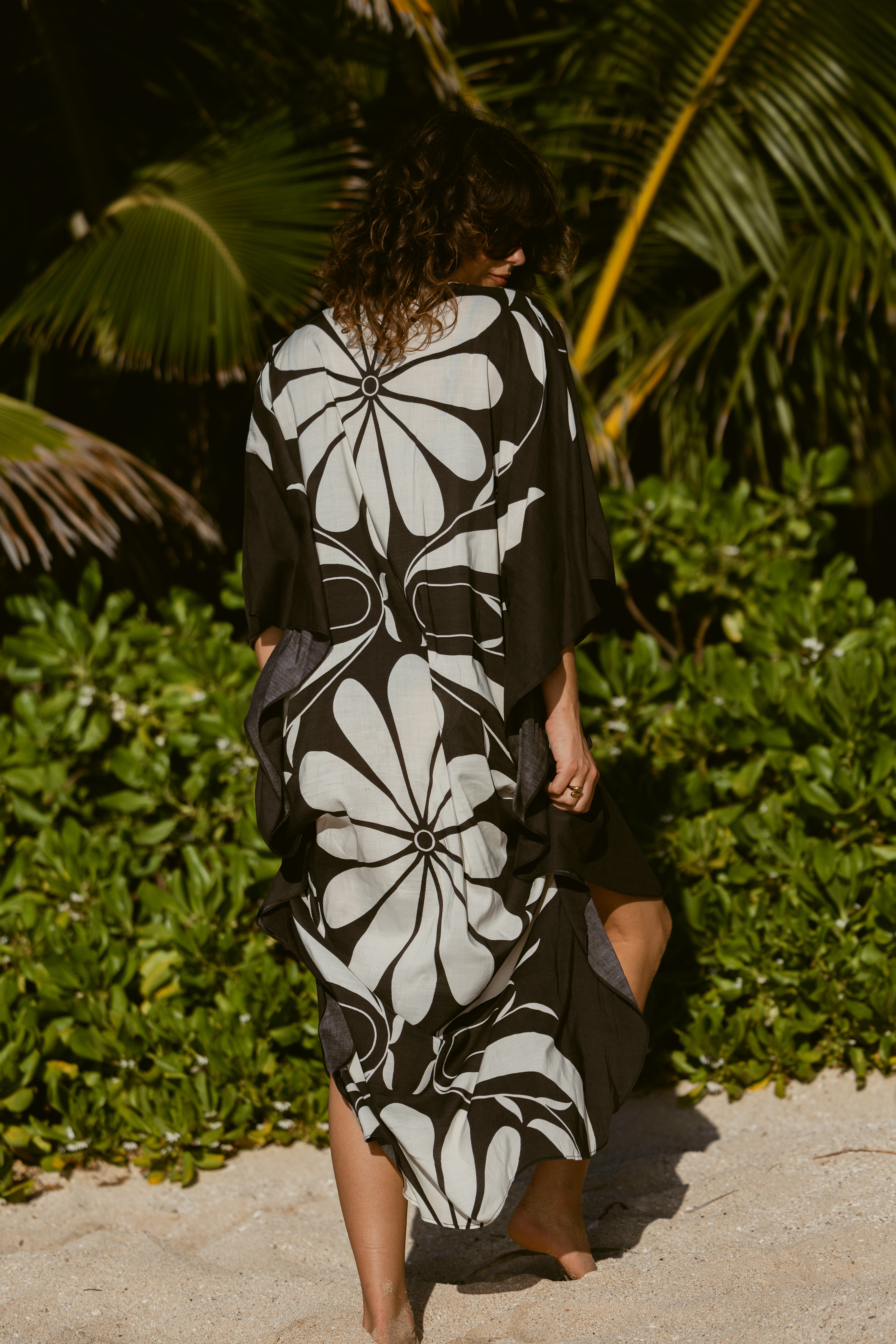 Wearing The Tropea Kaftan - Albatross by Dandy Del Mar, a person stands on a sandy beach with lush green foliage and palm leaves in the background.