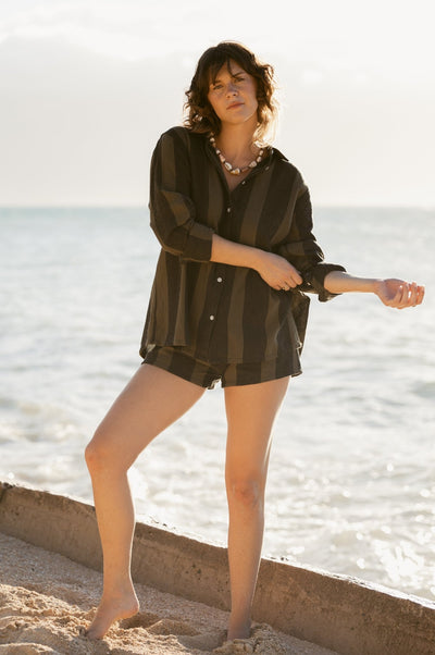 A person stands barefoot on the beach near the shoreline, wearing Dandy Del Mar’s Sevilla Shorts in Onyx and a loose, dark striped shirt with rolled sleeves on a sunny day.