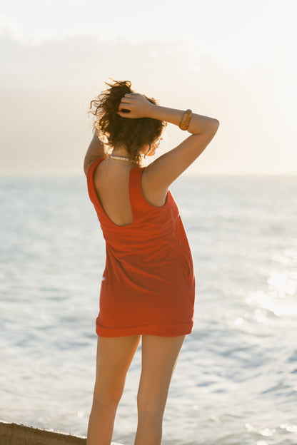 Wearing The Cyprus Dress by Dandy Del Mar, a person stands by the water facing the ocean, hands raised to adjust their hair.