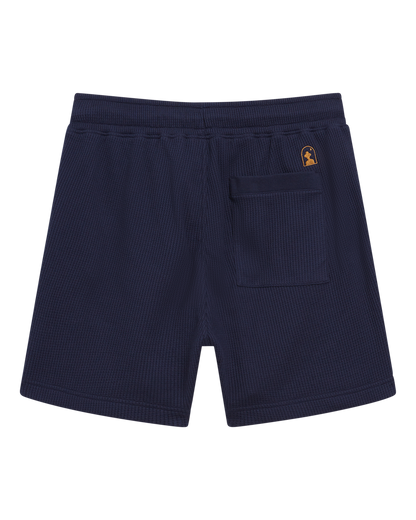 The Cannes Short - Luxe Navy by Dandy Del Mar is crafted from waffle knit, featuring an elastic waistband, a single back pocket, and a small gold logo patch above the pocket.