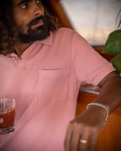 A man in the Dandy Del Mar Cannes Waffle Knit Polo Shirt in Spanish Rose holds a drink in his left hand, sitting indoors near a wooden surface and a green plant.