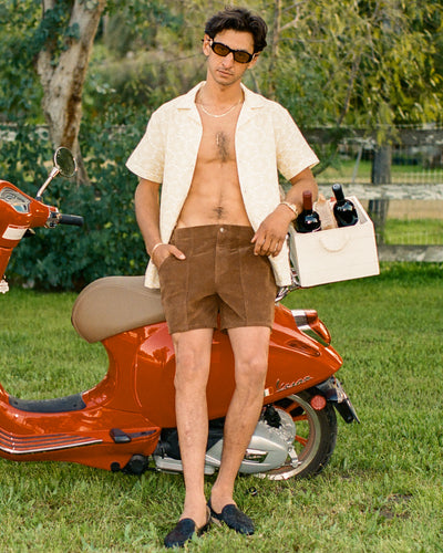 A man in sunglasses, an open short-sleeve shirt, and Dandy Del Mar’s The Corsica Corduroy Shorts in Tobacco stands on grass by a red scooter, holding a tote bag with wine bottles.