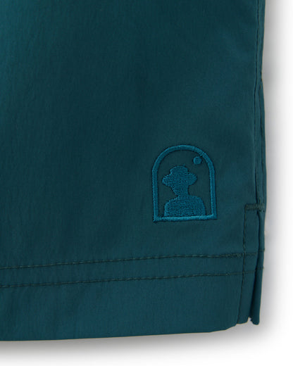 A detailed dark teal nylon spandex fabric features a small embroidered logo showing a head silhouette inside an arched frame, ideal for The Ventura Volley Short - Acqua by Dandy Del Mar, complete with an elastic waist.