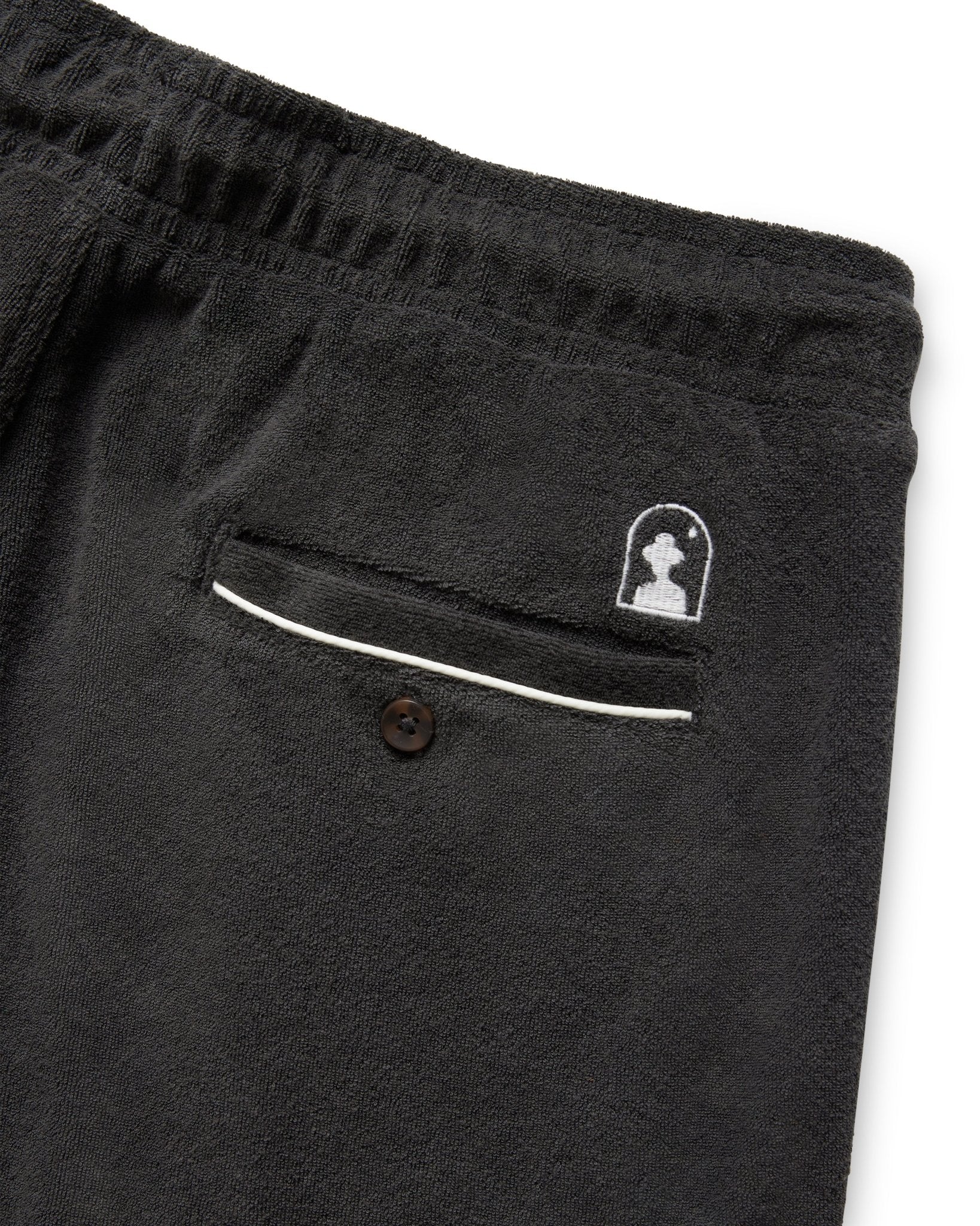 Close-up of The Gaucho Terry Cloth Shorts - Onyx by Dandy Del Mar, featuring black terry cloth, elastic waistband, white trim, a back pocket with button, and a small embroidered patch above the pocket for a classic look.